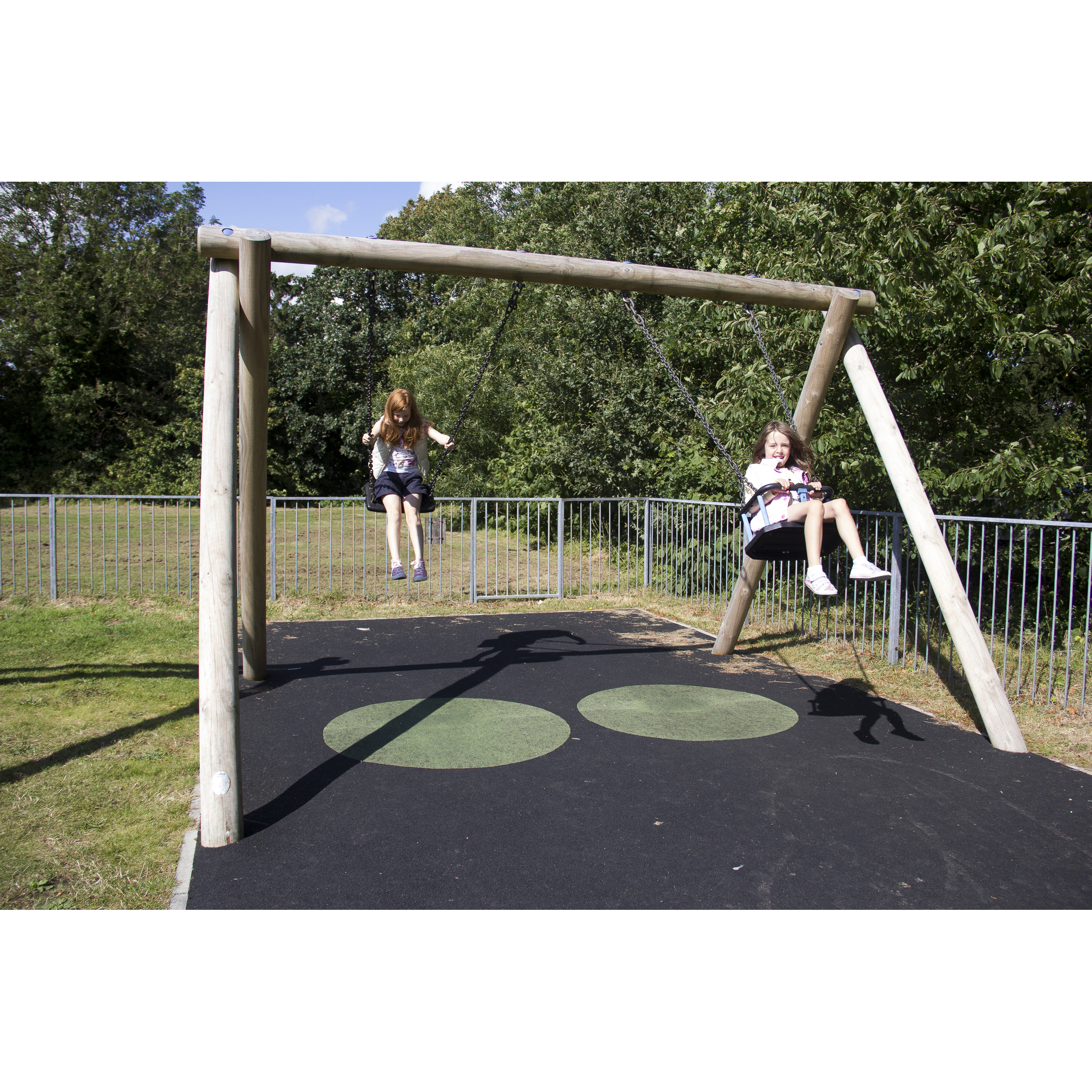 Two children are sitting on swings in a playground, with trees and a metal fence in the background on a sunny day.