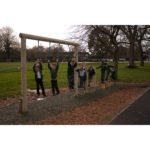 A group of children in school uniforms play on a wooden balance beam and climbing ropes in an outdoor playground.