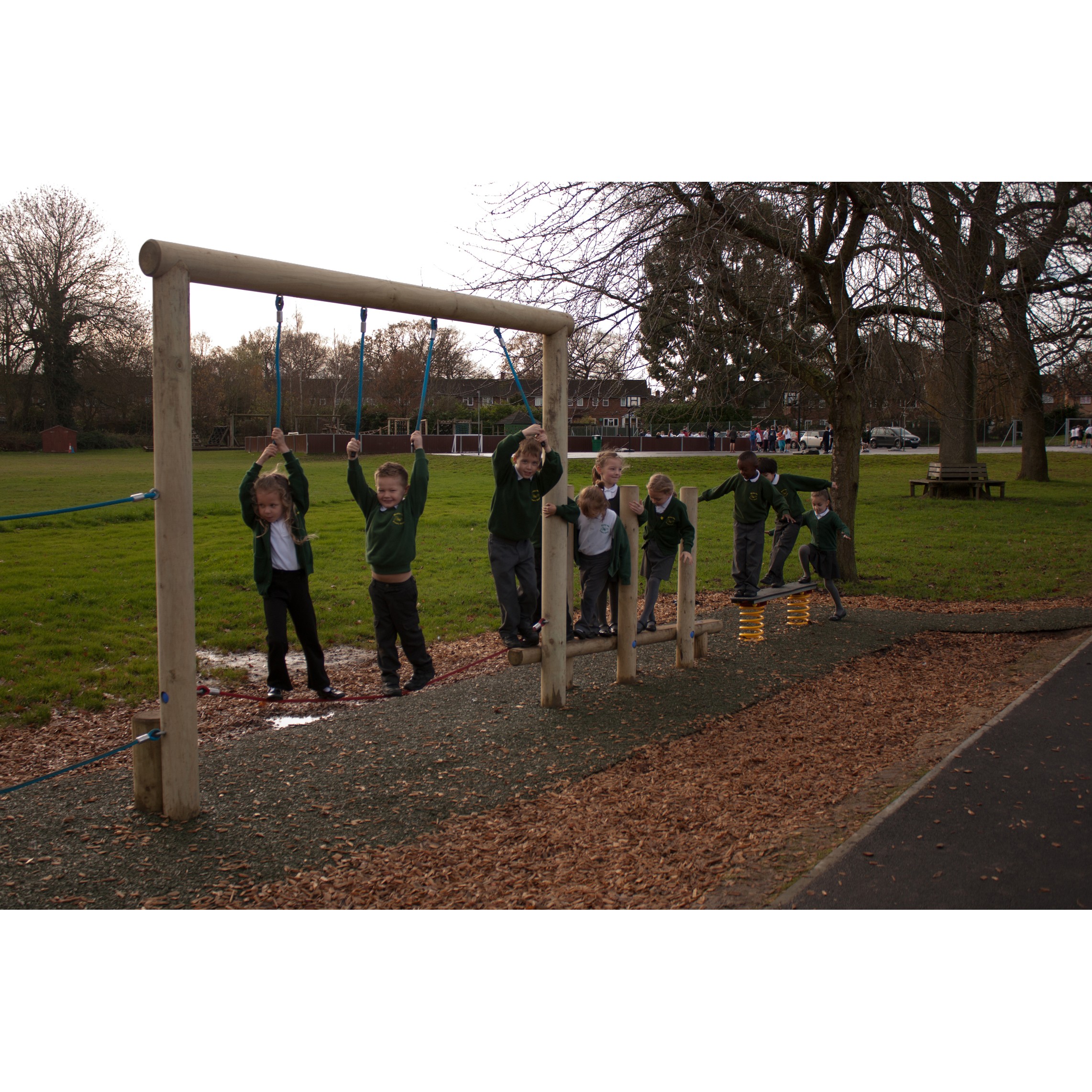 A group of children in school uniforms play on a wooden balance beam and climbing ropes in an outdoor playground.