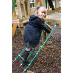 A young girl wearing a coat and boots climbs a rope ladder on a playground structure, smiling at the camera.