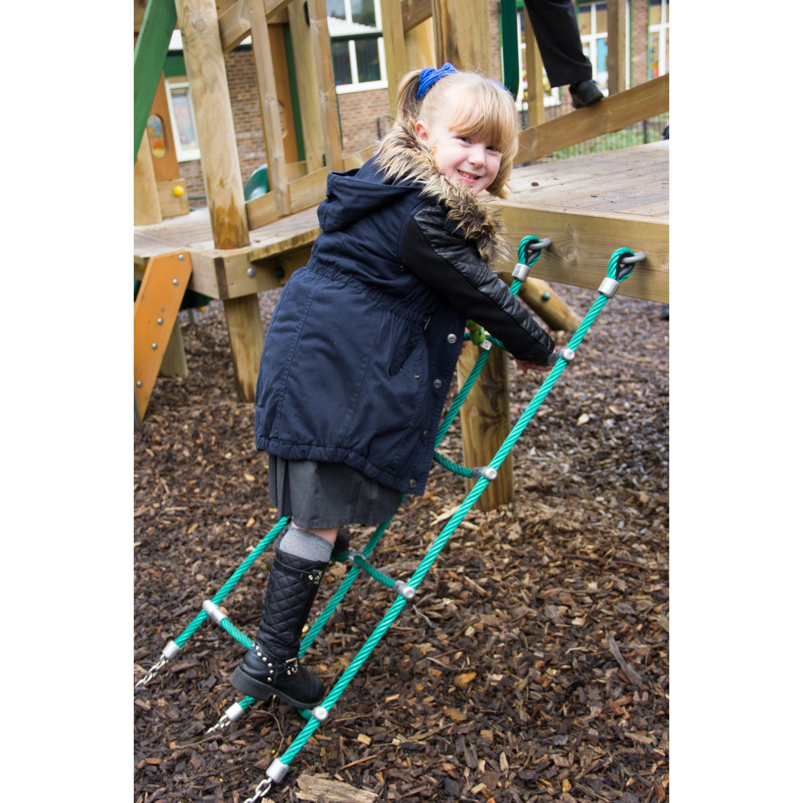 A young girl wearing a coat and boots climbs a rope ladder on a playground structure, smiling at the camera.