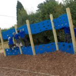 Children in school uniforms climb on a blue outdoor play wall with holes, surrounded by wood chippings and greenery.