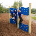 Four children in blue school uniforms climb on a blue outdoor climbing wall structure in a playground with woodchip ground covering.