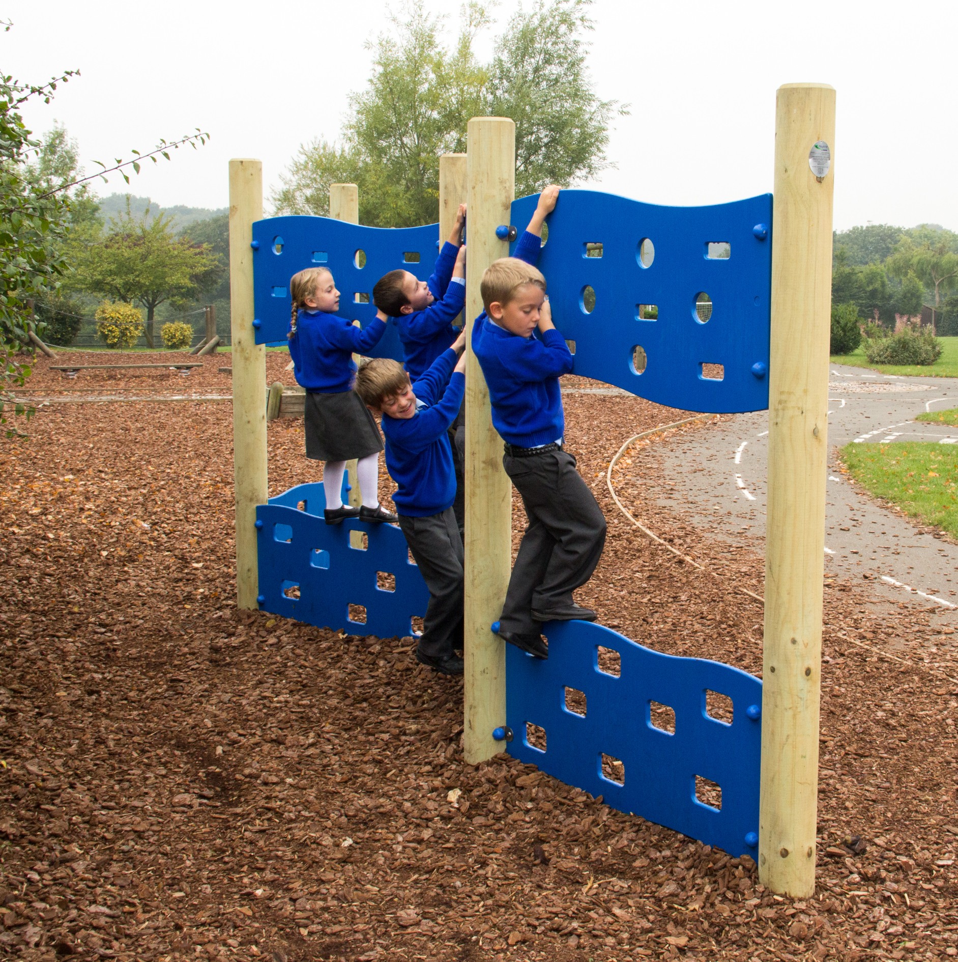 Four children in blue school uniforms climb on a blue outdoor climbing wall structure in a playground with woodchip ground covering.