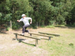 A person in a white shirt and cap jumps over a wooden exercise bar outdoors near a fence and trees.