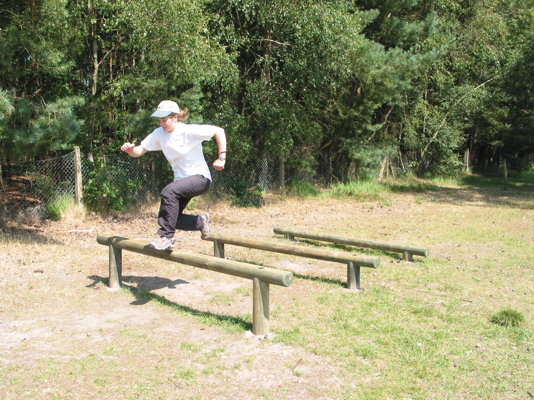 A person in a white shirt and cap jumps over a wooden exercise bar outdoors near a fence and trees.