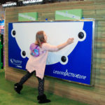 A girl touches a blue and white interactive wall panel labelled Learning ACTIVzone in an indoor play area.