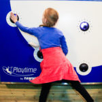 A young girl in a blue top and red skirt interacts with a blue and white wall panel featuring black buttons and the Playtime by Fawn logo.