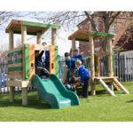 Several children in school uniforms play on a wooden playground structure with a green slide, set on artificial grass near a brick building and trees.