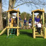 A group of children in school uniforms play on a wooden playground structure with rope bridges and ladders on a grassy area.