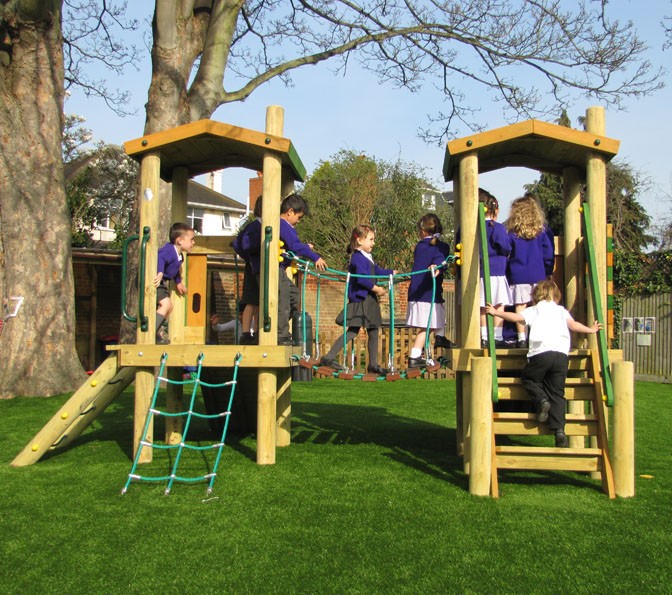 A group of children in school uniforms play on a wooden playground structure with rope bridges and ladders on a grassy area.