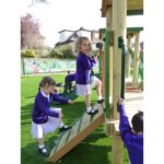 Two young girls in school uniforms climb a wooden playground structure whilst other children play nearby on artificial grass.