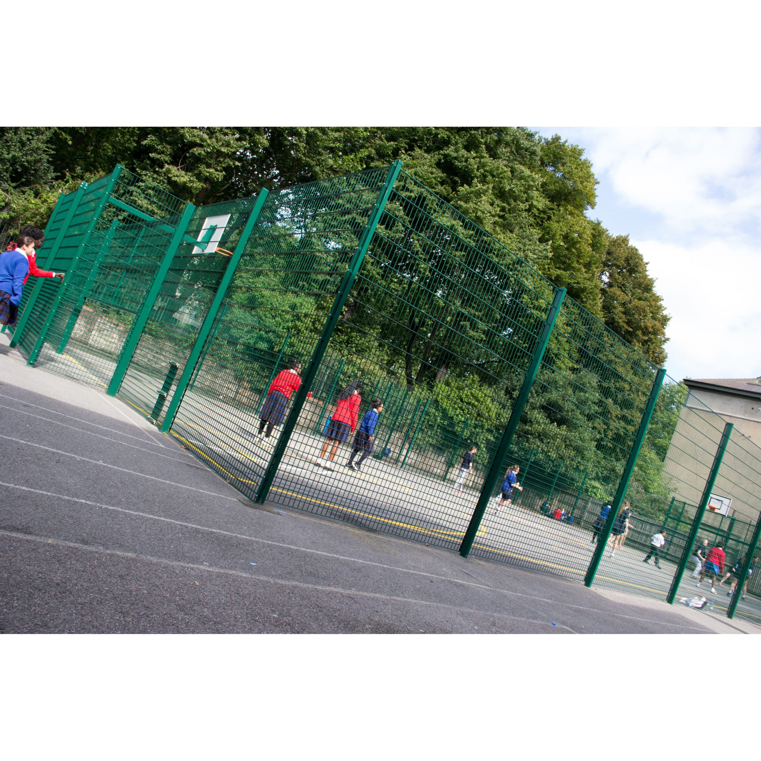 Several people play basketball on an outdoor Multi-Use Games Area surrounded by a tall green fence, with trees and buildings in the background.