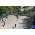 A group of people play basketball on an outdoor Multi-Use Games Area surrounded by green fencing, with spectators watching from the side.