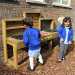 Three young children in blue uniforms play at an outdoor wooden mud kitchen set against a brick wall, surrounded by wood chippings.