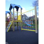 The Meteor Tower 900mm outdoor playground structure features climbing nets, a ladder, and a slide, all set on a black rubber surface with houses and trees in the background.