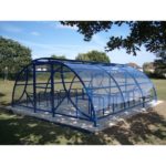 A blue metal and clear plastic bike shelter with bike racks underneath, located on a concrete slab in an open grassy area with trees and houses in the background.