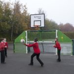 Four children in red jumpers play catch with a ball on a playground in front of a green and white basketball hoop and fence.