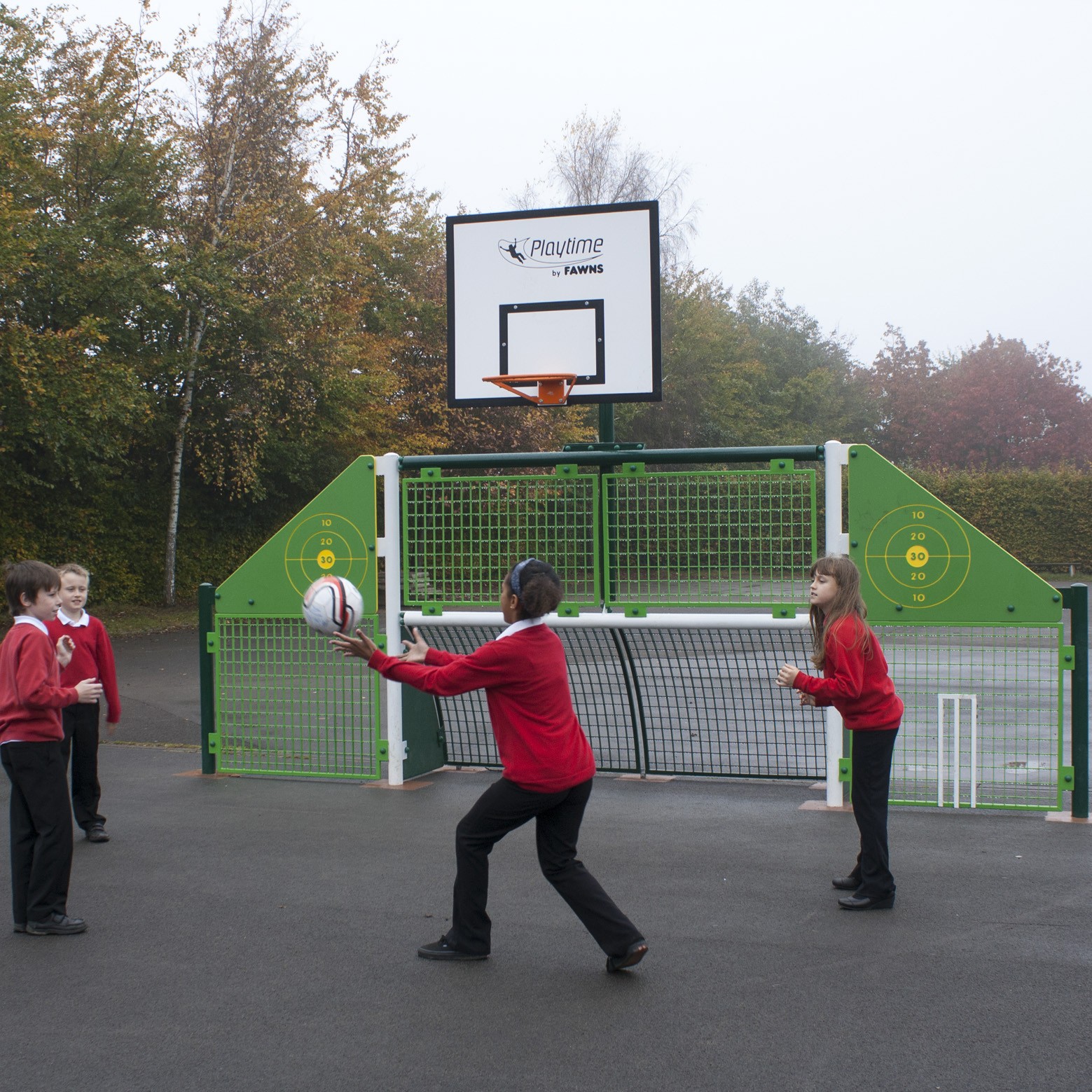 Four children in red jumpers play catch with a ball on a playground in front of a green and white basketball hoop and fence.