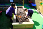 Two young children in school uniforms work in a raised 35cm log planter, one using a small spade and the other digging with hands, enjoying gardening in an outdoor play area.