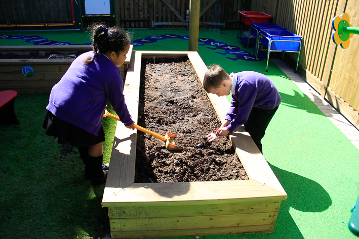 Two young children in school uniforms work in a raised 35cm log planter, one using a small spade and the other digging with hands, enjoying gardening in an outdoor play area.