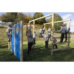 Children in school uniforms play on a wooden climbing frame and Orbiter Space Frame outdoors on a grassy area, with trees and a building in the background.