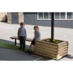 Two young children in grey sweatshirts sit and stand by a wooden planter with yellow flowers in an outdoor schoolyard area.
