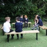 Six children in school uniforms sit and talk on wooden benches outdoors, surrounded by greenery and trees.