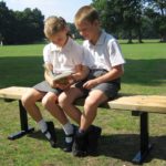 Two schoolchildren in uniform sit on a bench outdoors, reading a book together on a sunny day in a park or playground.