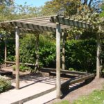 A wooden garden pergola stands on a paved area surrounded by green bushes and trees, with some climbing plants growing on the structure.
