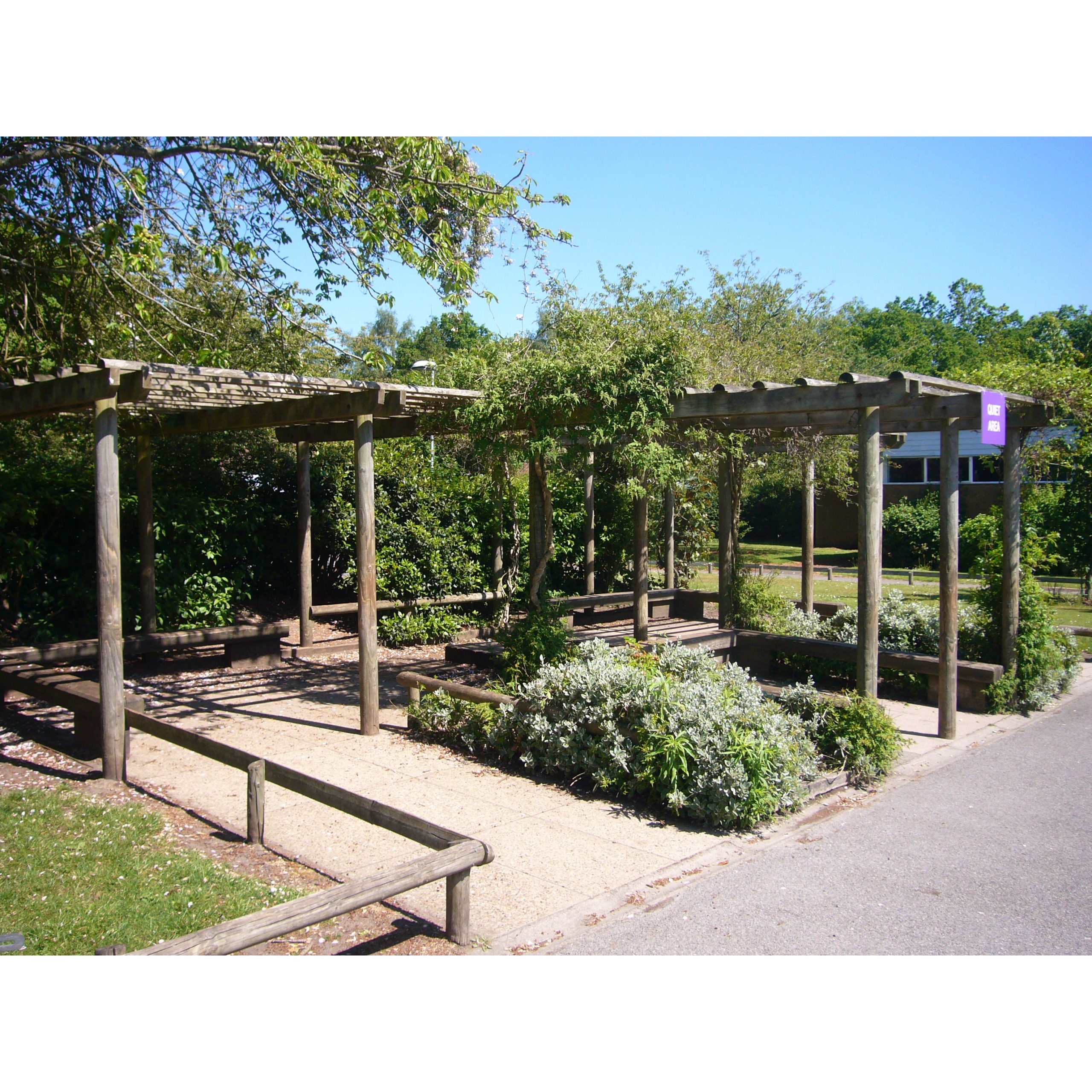 Wooden pergolas with benches and greenery underneath, situated in a landscaped outdoor area on a sunny day.