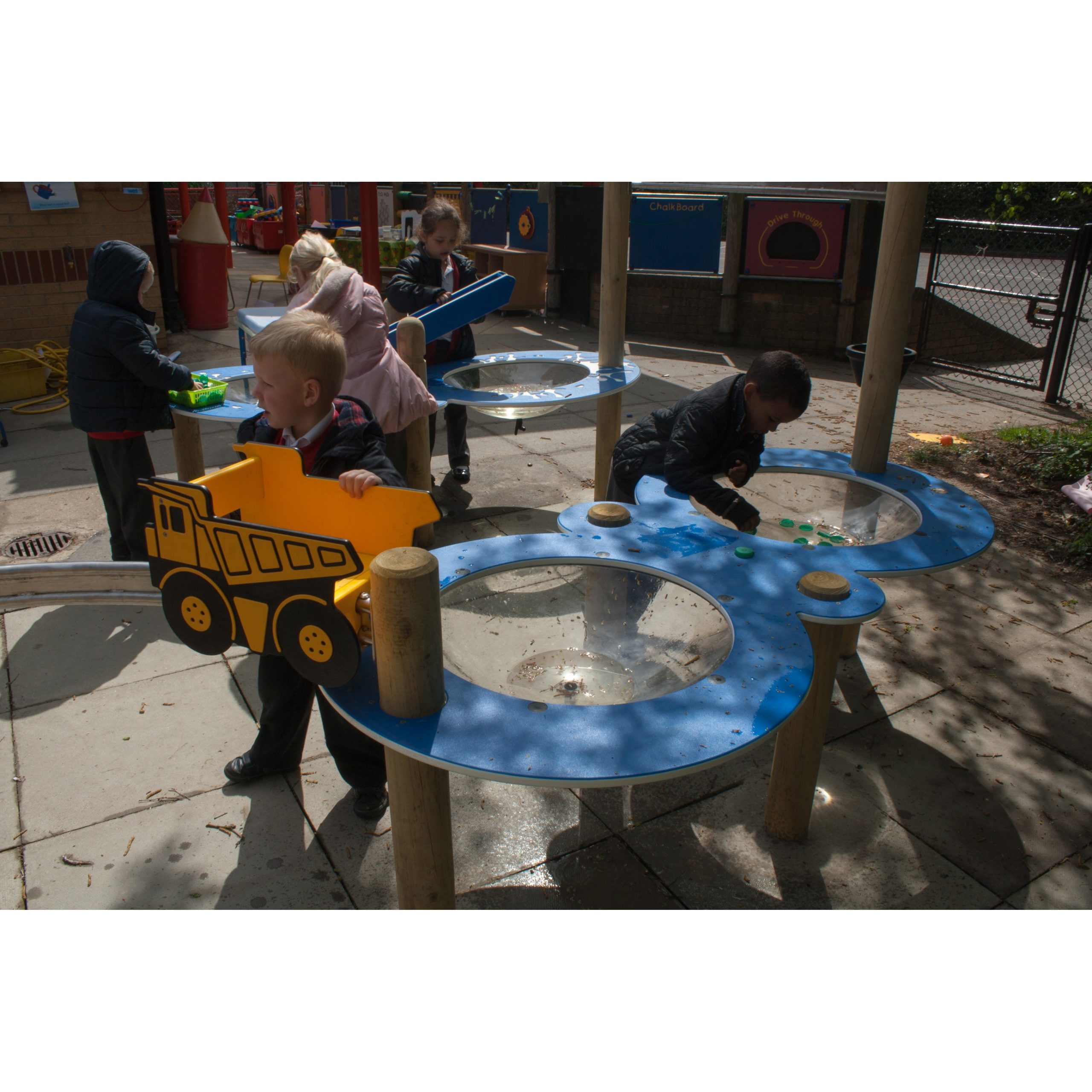 Several young children play with water tables and toy lorries outdoors on a sunny day.