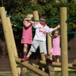 Three children play on a wooden climbing frame outdoors; one wears a white sports shirt, while the others in pink dresses look as though they’re balancing in a game of Pick Up Sticks 1.