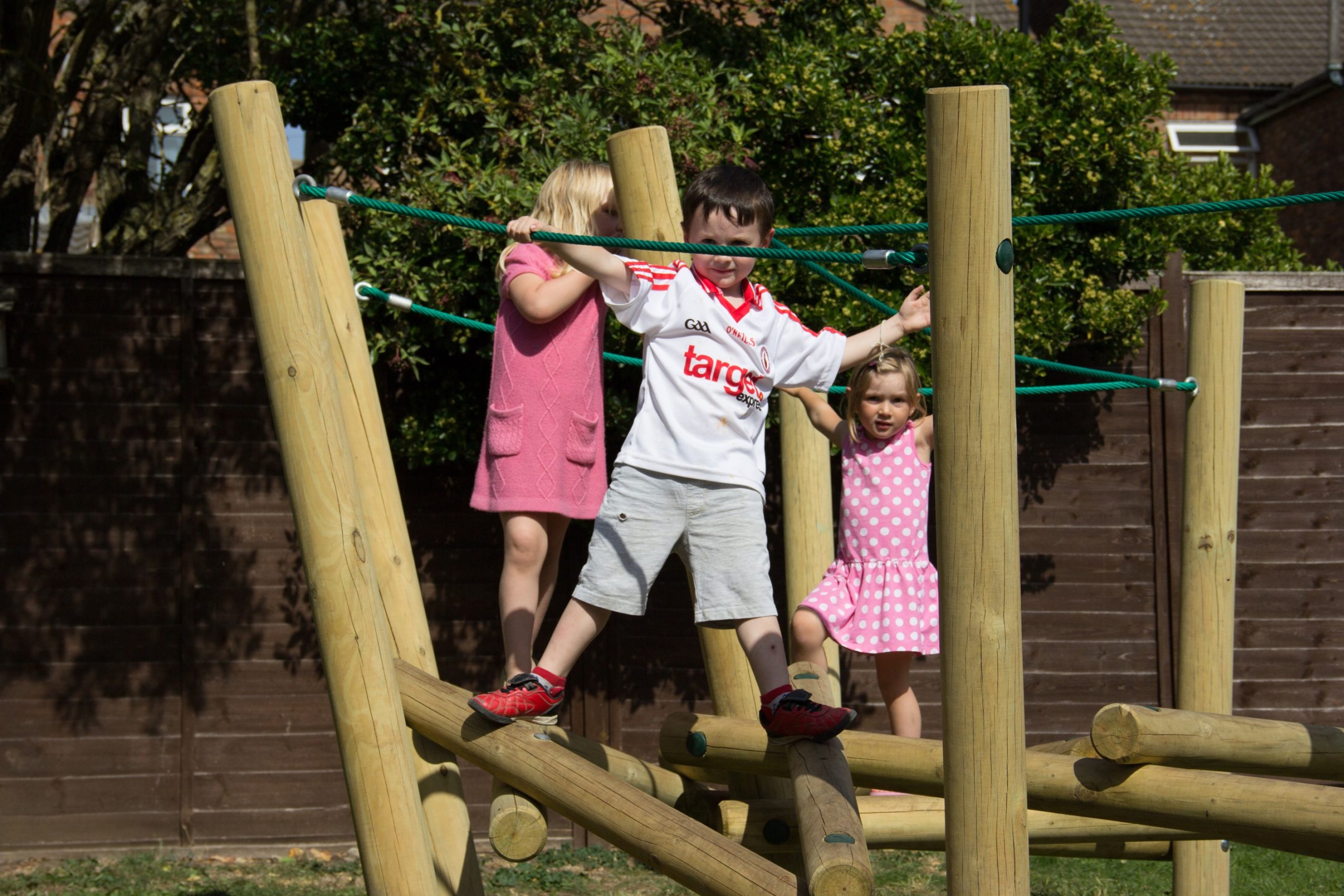 Three children play on a wooden climbing frame outdoors; one wears a white sports shirt, while the others in pink dresses look as though they’re balancing in a game of Pick Up Sticks 1.