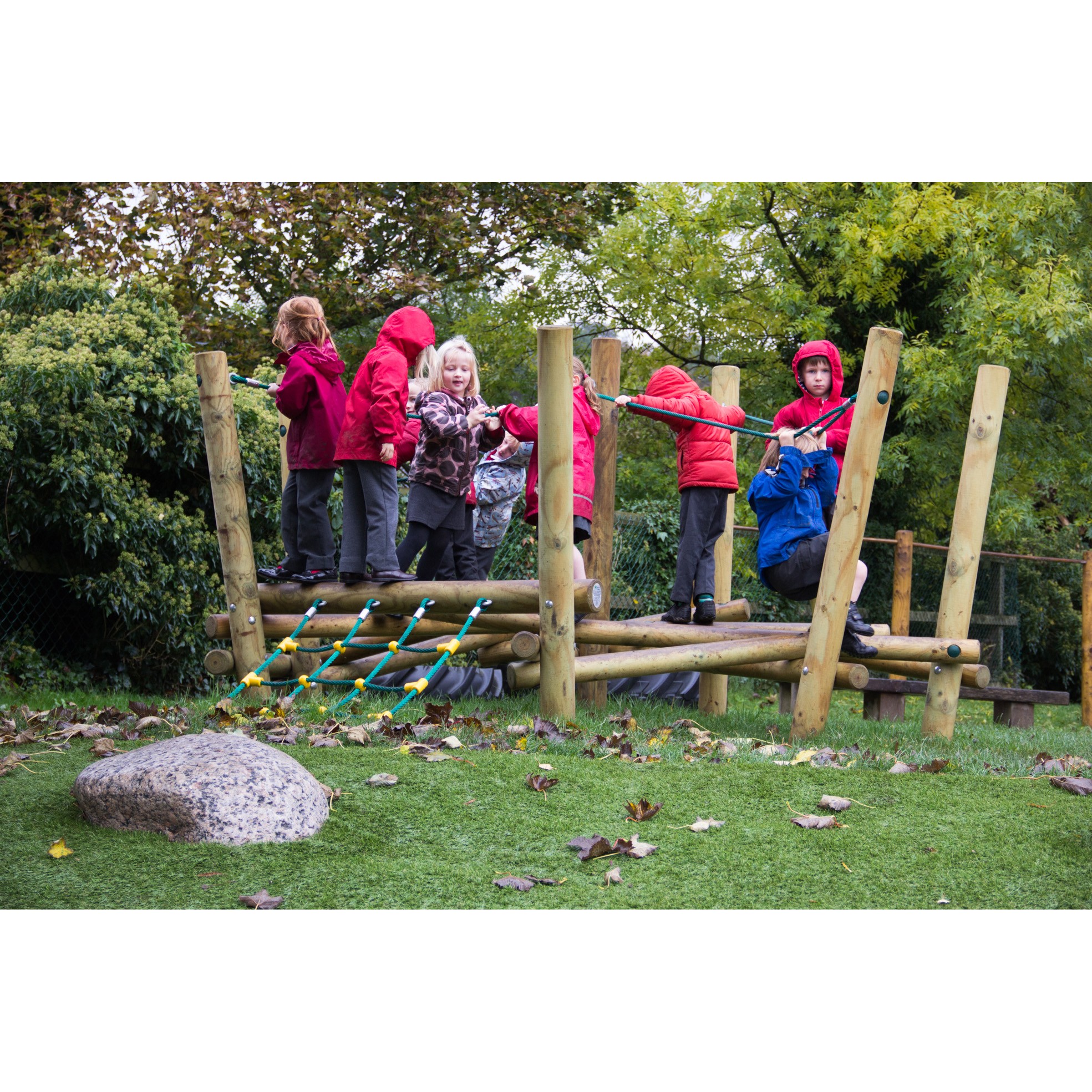 Several children in jackets and raincoats play on the Pick Up Sticks 1 wooden climbing frame outdoors, surrounded by grass, leaves, and trees.