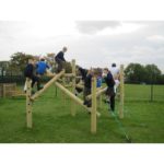 Children in school uniforms climb and play on a wooden playground structure outdoors on a grassy field.