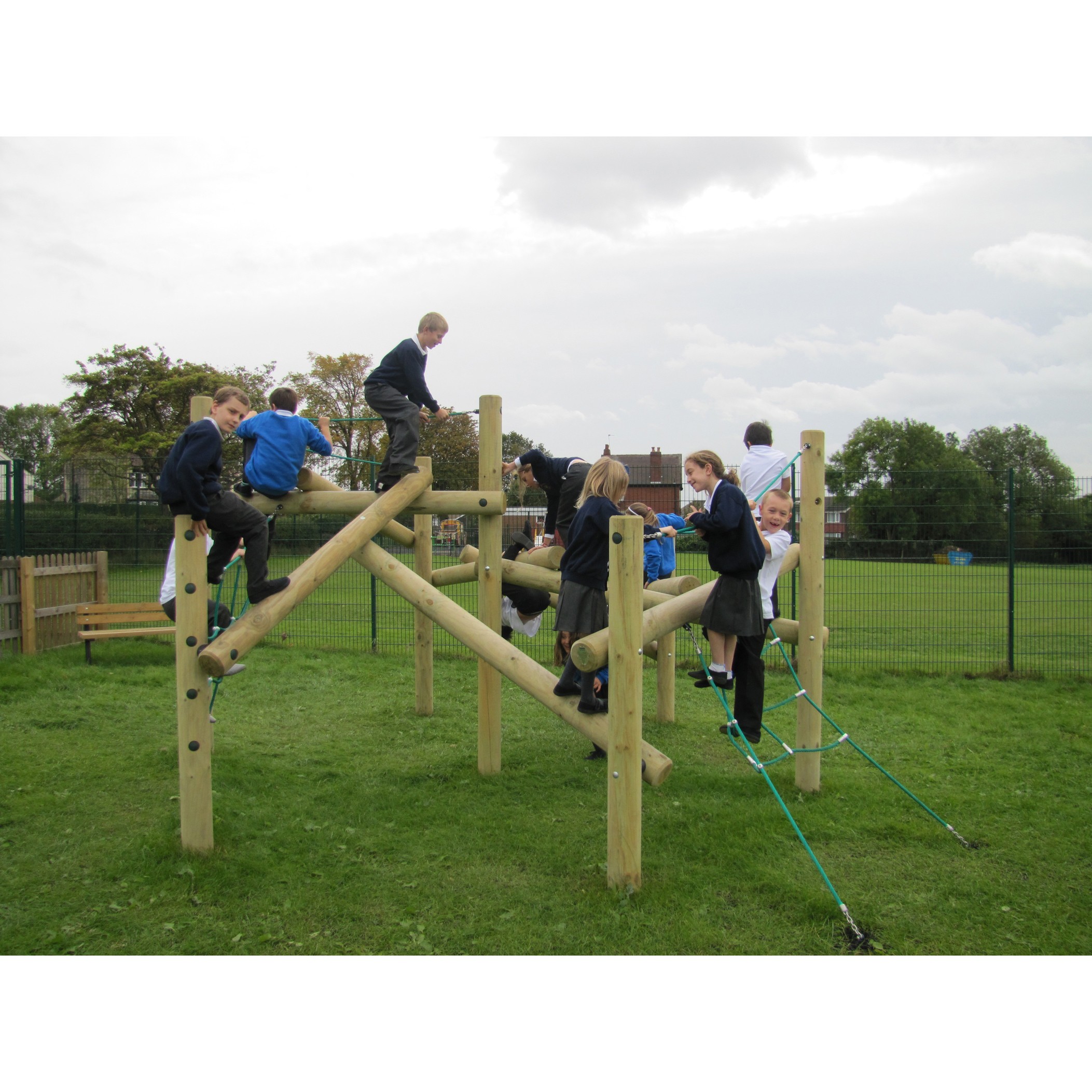 Children in school uniforms climb and play on a wooden playground structure outdoors on a grassy field.