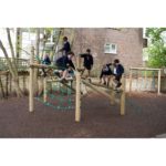 Six children in school uniforms play on a wooden climbing frame with rope nets in an outdoor playground area surrounded by trees and a brick building.