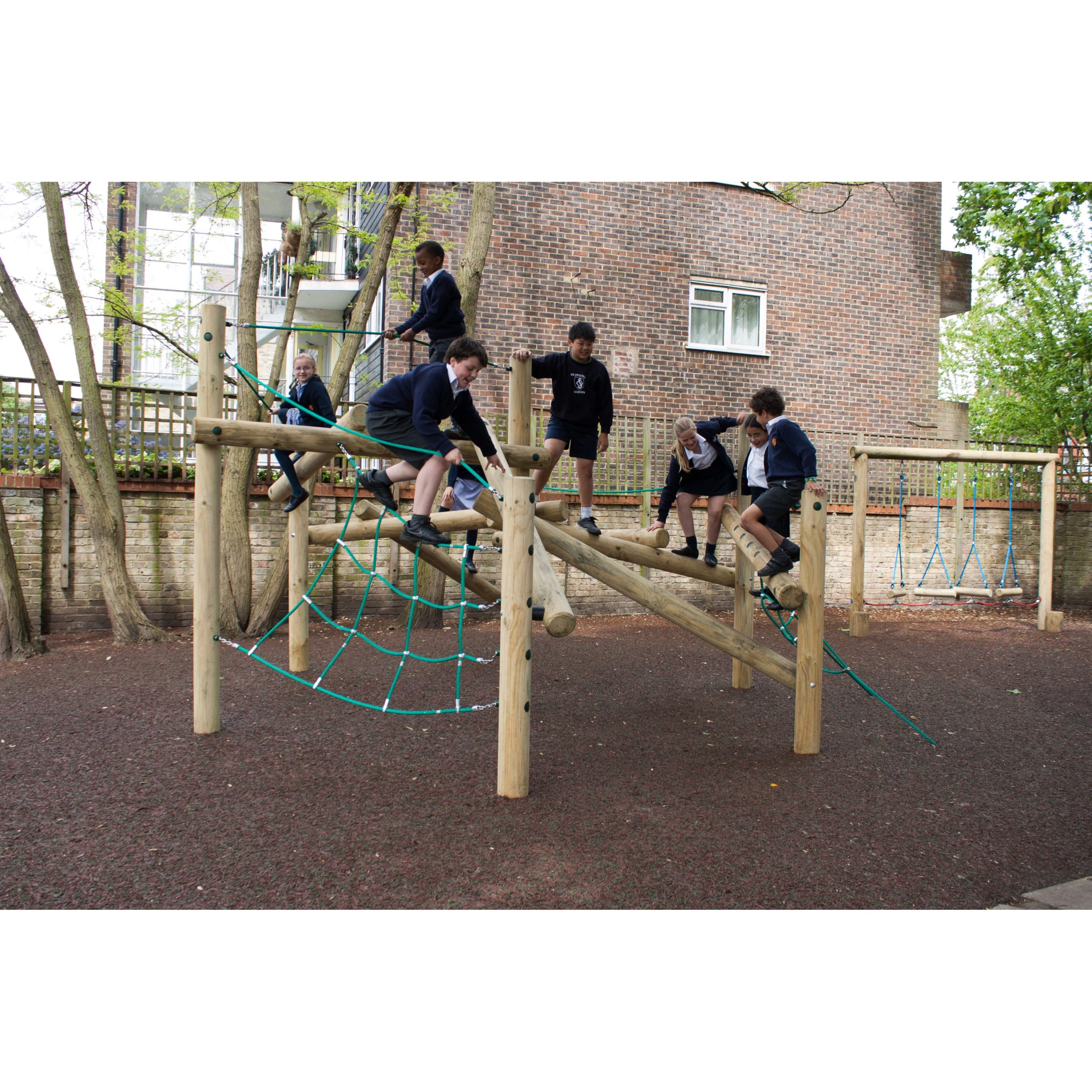 Six children in school uniforms play on a wooden climbing frame with rope nets in an outdoor playground area surrounded by trees and a brick building.