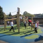Children play and climb on a wooden playground structure with ropes on a padded surface in an outdoor schoolyard.