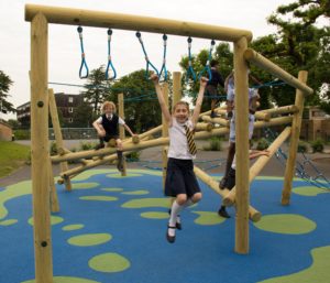 Children in school uniforms play on a wooden climbing frame and ropes at an outdoor playground with blue and green safety flooring.