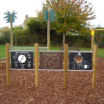 Children’s playground panels with pirate-themed activities, including a directional map, a treasure map, and a cut-out pirate face panel, on wood chip ground with trees in the background.