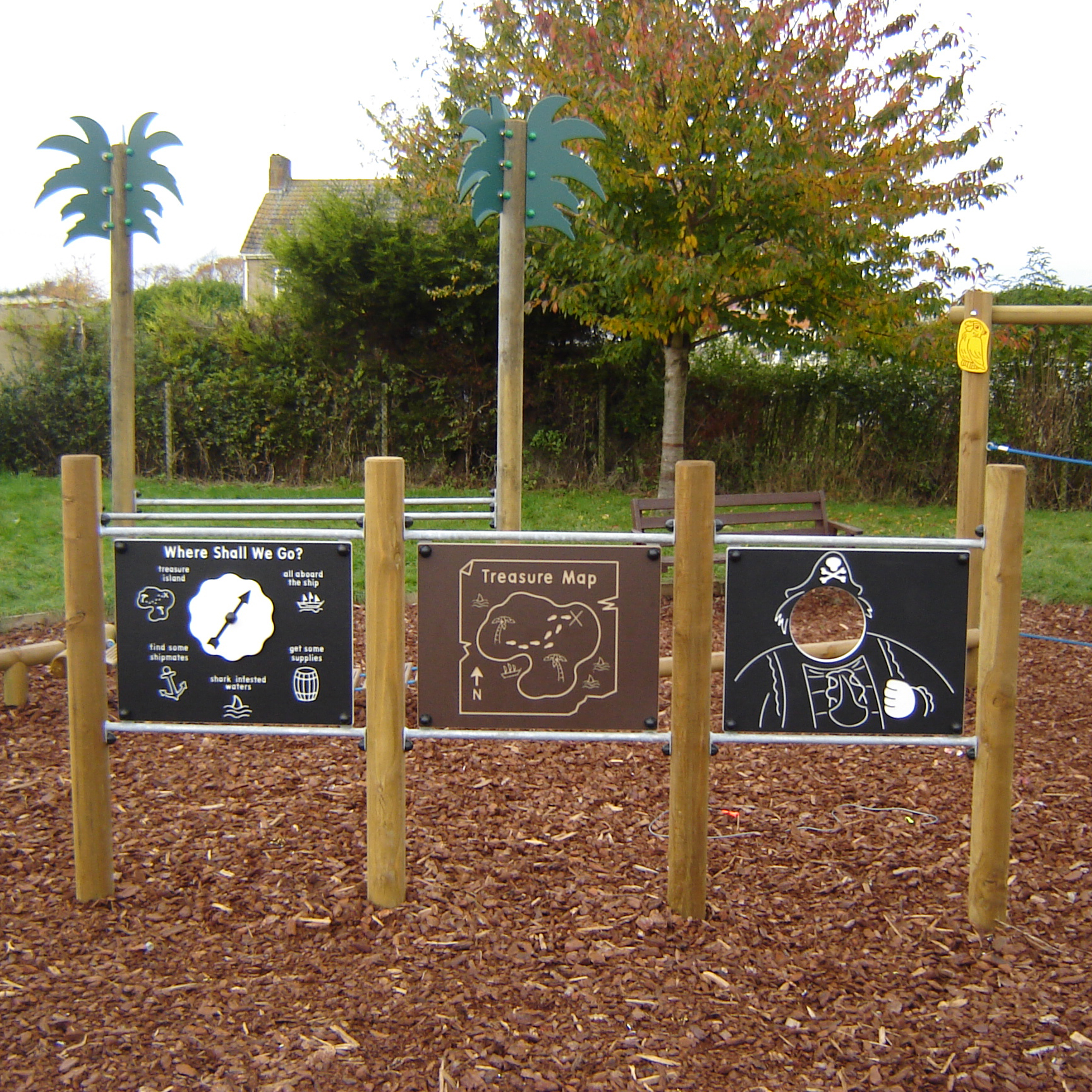 Children’s playground panels with pirate-themed activities, including a directional map, a treasure map, and a cut-out pirate face panel, on wood chip ground with trees in the background.