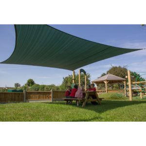 Children sit at a picnic table under a large green sunshade in a grassy outdoor play area, with climbing equipment and a gazebo nearby.