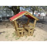 A small wooden playhouse with a red roof and built-in benches, situated on a sandy area under a tree in an outdoor playground.
