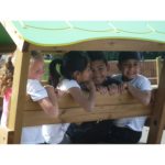 Four children sit closely together on a wooden playground bench under a green canopy, smiling and looking at each other.