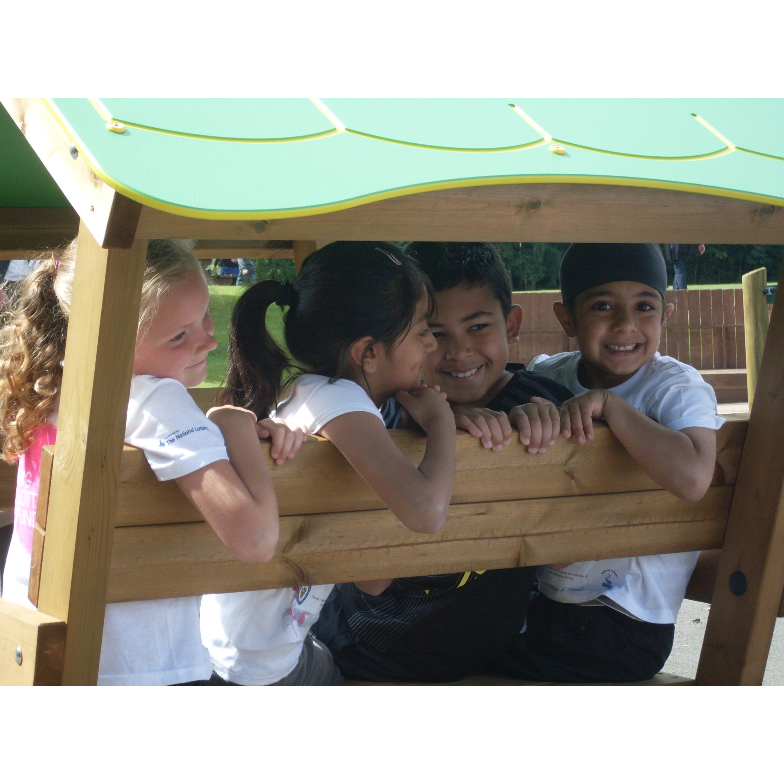 Four children sit closely together on a wooden playground bench under a green canopy, smiling and looking at each other.