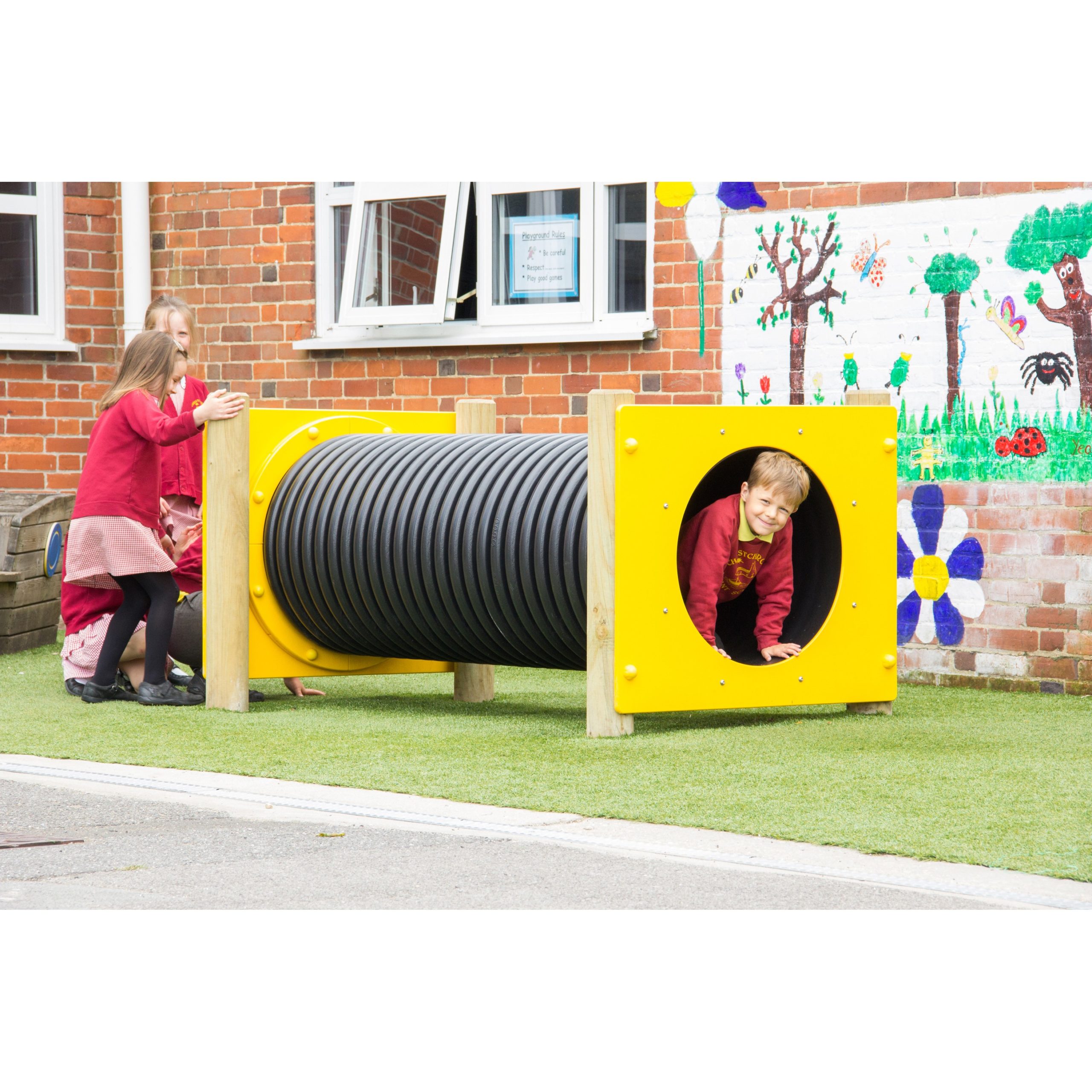 Three children play outside; one crawls through a black plastic tunnel with yellow ends whilst two others wait nearby. A mural with flowers and insects is visible on the brick wall behind them.