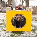 Five children play outside on a snowy day; one child crawls through a yellow playground tunnel whilst four others stand behind it.