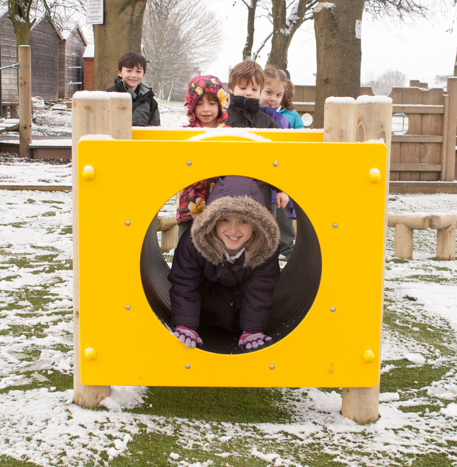 Five children play outside on a snowy day; one child crawls through a yellow playground tunnel whilst four others stand behind it.